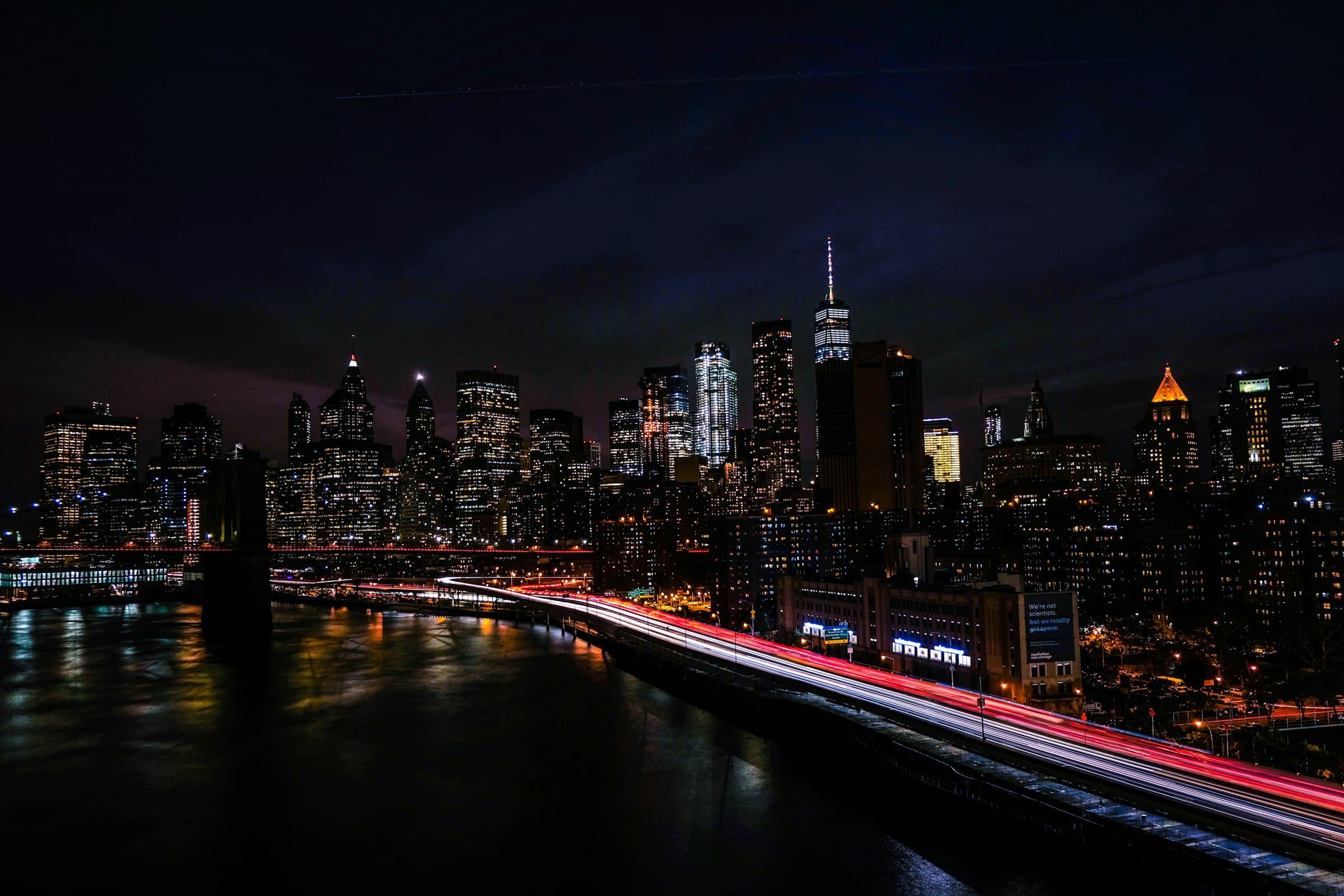 The Lower Manhattan skyline at night, with light trails along the riverside highway
